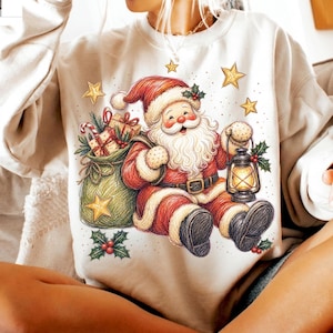 K&ouml;nnte beinhalten: Ein cremefarbenes Sweatshirt mit einer grafischen Illustration des Weihnachtsmanns in einem roten Anzug mit wei&szlig;em Besatz, der eine leuchtende Laterne h&auml;lt. Neben dem Weihnachtsmann steht ein gr&uuml;ner Sack voller Geschenke, umgeben von goldenen Sternen und Stechpalmen.