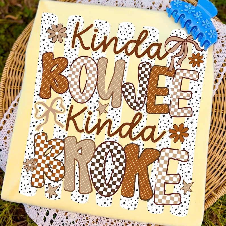 Pode incluir: Uma t-shirt amarelo-claro com o texto castanho Kinda BOUJEE Kinda BROKE. O design inclui padr&otilde;es axadrezados castanhos e bege, pontos, pequenas flores, estrelas e la&ccedil;os. Um gancho de cabelo de pl&aacute;stico azul brilhante repousa no canto superior.