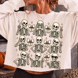 May include: A cream-colored long-sleeve sweatshirt featuring a graphic print of nine skeletons in various poses, including waving, making peace signs, and wearing sunglasses, interspersed with small star icons and several decorative bows.