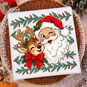 May include: A white square fabric item featuring a colorful graphic of Santa Claus and a reindeer. Santa wears a red hat and has a white beard. The reindeer has brown fur and a large red bow. Green pine branches and red lights frame the design.