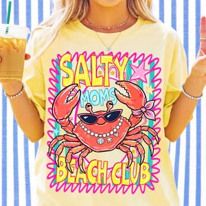 May include: A pale yellow t-shirt featuring a graphic of a red crab wearing black sunglasses and a pearl necklace. The text reads SALTY MOMS BEACH CLUB in yellow letters. The design includes pink star accents and a bright pink wavy border.