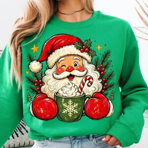 May include: A bright green crewneck sweatshirt featuring a graphic of Santa Claus wearing a red hat and red mittens, winking while holding a green mug filled with whipped cream and a candy cane, surrounded by pine branches, holly, and stars.