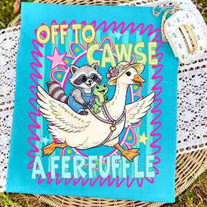 May include: A teal-colored graphic featuring a cartoon goose wearing a hat, carrying a raccoon and a frog. The text "OFF TO CAWSE A FERFUFFLE" is displayed in a playful font, surrounded by pink squiggly lines and stars. The design is on a square fabric.