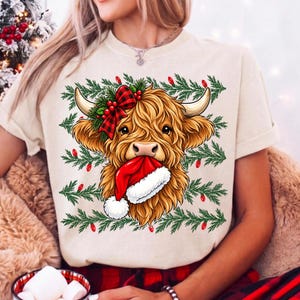May include: A cream-colored t-shirt featuring a graphic of a fluffy Highland cow with a red and black buffalo plaid bow on its head and a red Santa hat in its mouth, set against a background of green pine branches and small red Christmas lights.