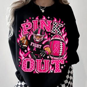 K&ouml;nnte beinhalten: Ein schwarzes Sweatshirt mit einem Grafikdruck und dem Text PINK OUT. Das Design zeigt einen Footballspieler in schwarz-rosa Uniform, einen rosa Football mit einem Schleifensymbol sowie rosa Flammen, Sterne und Punkte-Muster.
