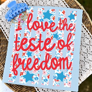 Op de afbeelding: Een lichtblauw vierkant bord met verticale strepen met een bloemenpatroon in rood, roze en blauw. De rode tekst luidt I love the teste of freedom. Vier blauwe sterren zijn over het bord verspreid. Een blauwe bloem-haarklem en kralenarmbanden.