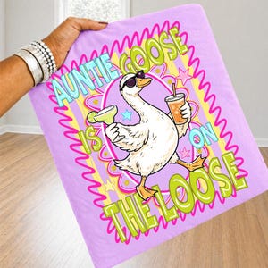K&ouml;nnte beinhalten: Lavendelfarbene Decke mit dem Aufdruck &bdquo;Auntie Goose is on the Loose&ldquo;. Die Grafik zeigt eine Cartoon-Gans mit Sonnenbrille, Margarita und Getr&auml;nk. Text in Gr&uuml;n, Gelb und Blau.