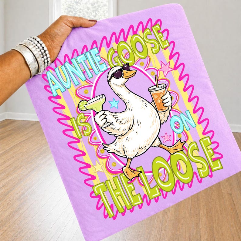 K&ouml;nnte beinhalten: Lavendelfarbene Decke mit dem Aufdruck &bdquo;Auntie Goose is on the Loose&ldquo;. Die Grafik zeigt eine Cartoon-Gans mit Sonnenbrille, Margarita und Getr&auml;nk. Text in Gr&uuml;n, Gelb und Blau.