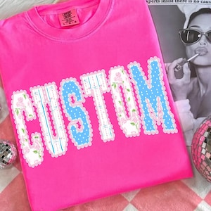 May include: Bright pink t-shirt with the word "CUSTOM" in large, decorative letters. The letters have a blue polka dot and floral design. The shirt is made of a soft material and has a crew neck.