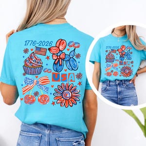 May include: A bright turquoise t-shirt featuring a patriotic graphic print with the text 1776-2026 and USA. The design includes a balloon dog, a cupcake, a bow, cherries, and a flower, all rendered in red, white, and blue patriotic colours.