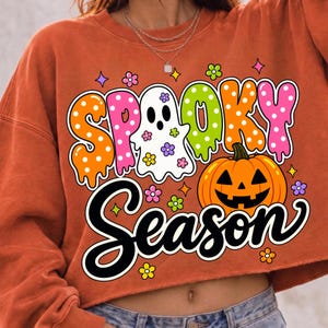 May include: An orange cropped sweatshirt featuring a graphic print with the text "SPOOKY SEASON". The design includes a white ghost, a jack-o'-lantern, and colorful flowers in shades of pink, green, and yellow against the orange fabric.