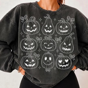 May include: A charcoal grey mineral-washed crewneck sweatshirt featuring a white line-art graphic print. The design displays nine distinct jack-o'-lantern pumpkins with varied facial expressions, interspersed with small star and bow icons.