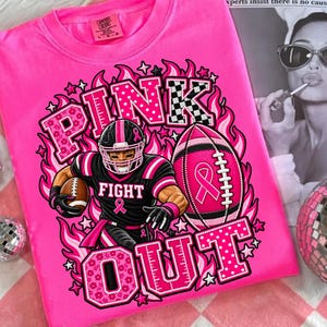 K&ouml;nnte beinhalten: Ein leuchtend pinkes T-Shirt mit einer Grafik eines Footballspielers in schwarz-pinker Uniform, der einen Football h&auml;lt. Das Design umfasst einen pinken Football mit Schleife, Flammen, Sternen sowie den Text "PINK OUT" und "FIGHT" in Fettdruck.