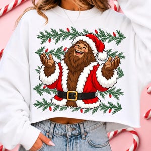 Puede incluir: Una sudadera blanca corta con una ilustraci&oacute;n gr&aacute;fica de un Bigfoot marr&oacute;n vestido con un traje de Pap&aacute; Noel rojo con ribetes de piel blanca, un cintur&oacute;n negro con hebilla dorada y un gorro de Navidad, rodeado de ramas de pino verdes y luces.