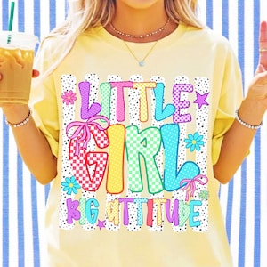 May include: A pale yellow t-shirt featuring a multi-colored graphic print with the text "LITTLE GIRL BIG ATTITUDE". The design includes vertical black dotted stripes, checkered patterns, and small flower and star icons in pink, blue, and yellow.