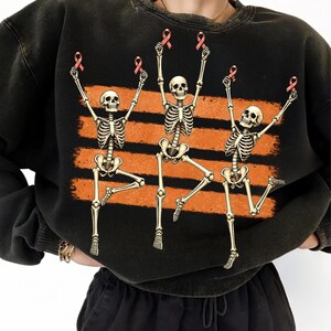 May include: A black crewneck sweatshirt featuring a graphic print of three dancing skeletons. Each skeleton is holding a small pink ribbon above its head. The skeletons are positioned over three horizontal, distressed orange brushstroke stripes.