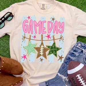 May include: A cream-colored short-sleeve t-shirt featuring a graphic print with the text "GAME DAY" in pink letters. The design includes a clothesline with three star-shaped ornaments, one resembling a football, against light blue vertical stripes.