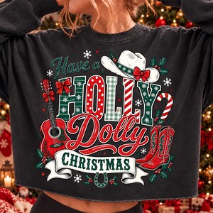 May include: A charcoal gray cropped sweatshirt featuring a festive graphic with the text Have a HOLLY Dolly CHRISTMAS. The design includes a red acoustic guitar, a white cowboy hat with a red bow, red cowboy boots, holly leaves, and snowflakes.