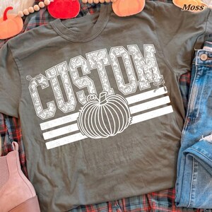 May include: A dark olive green short-sleeve t-shirt featuring the word CUSTOM in large white block letters filled with a small pumpkin pattern. Below the text is a large white line-art pumpkin graphic with three horizontal white stripes.