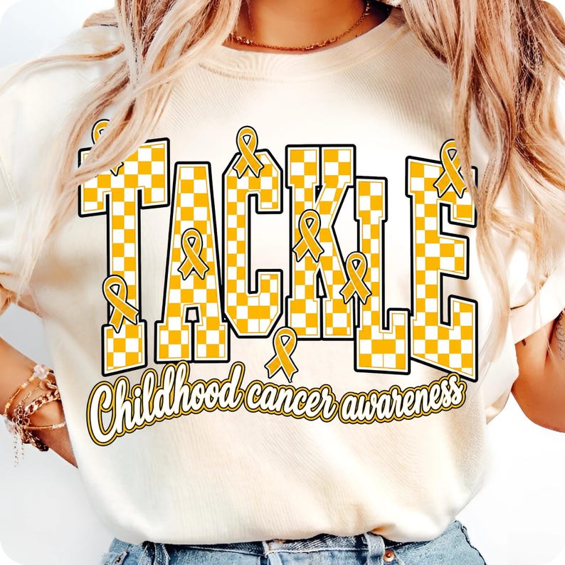 May include: A cream-colored t-shirt featuring the word TACKLE in large, yellow and white checkered block letters. Several yellow awareness ribbons are integrated into the letters. Below, the text Childhood cancer awareness is printed in yellow.