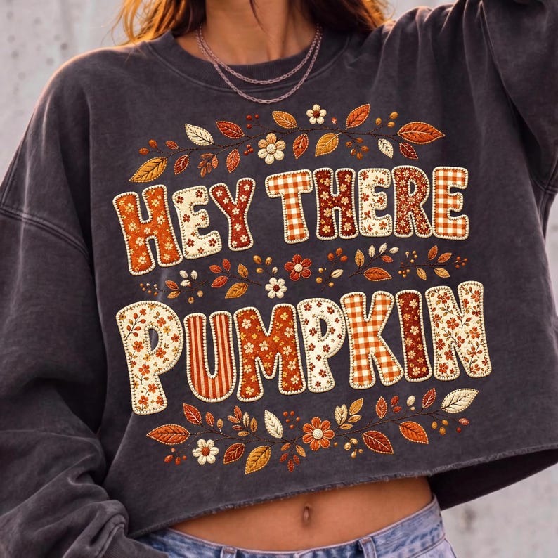 May include: A charcoal grey cropped sweatshirt featuring the text HEY THERE PUMPKIN in a patchwork style with various orange, brown, and cream floral and plaid patterns. The design is surrounded by embroidered autumn leaves and small flowers.