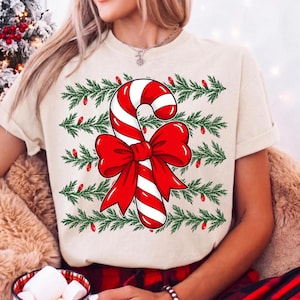 May include: A cream-colored short-sleeve t-shirt featuring a central graphic of a red and white striped candy cane tied with a large red bow. The design is layered over horizontal rows of green pine branches accented with small red Christmas lights.
