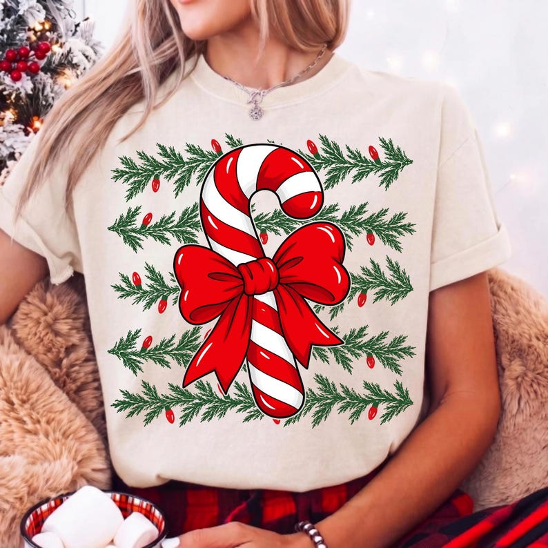 May include: A cream-colored short-sleeve t-shirt featuring a central graphic of a red and white striped candy cane tied with a large red bow. The design is layered over horizontal rows of green pine branches accented with small red Christmas lights.