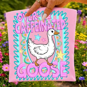 Pu&ograve; includere: Un quadrato di tessuto rosa chiaro con un'illustrazione centrale di un'oca bianca. Il testo recita OVER-CAFFEINATED GOOSE in lettere rosa in grassetto. Il design include un bordo smerlato turchese, strisce verticali azzurre e stelle gialle e rosa.