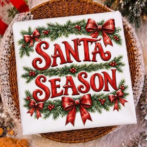 以下が含まれることがあります： 装飾的なフォントで赤い文字「SANTA SEASON」がプリントされた白い正方形の布地。デザインは緑の松の枝、赤いベリー、そして四隅と中央に配置された4つの赤いチェック柄のリボンで縁取られています。