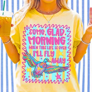 Pu&ograve; includere: Una t-shirt a maniche corte color giallo pallido con una grafica che riporta il testo SOME GLAD MORNING WHEN THIS LIFE IS OVER I'LL FLY AWAY. Il design include un'anatra volante colorata, stelle gialle e un bordo rosa ondulato su sfondo a righe azzurre.
