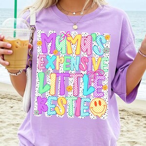 May include: A light purple t-shirt featuring a graphic print with the text Mama's Expensive Little Bestie. The design includes colorful checkered patterns, small flowers, pink bows, and a yellow smiley face, all set against vertical black dotted lines.