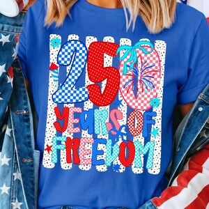 May include: A royal blue short-sleeve t-shirt featuring a graphic print with the text 250 YEARS OF FREEDOM. The design includes red, white, and blue checkered and polka-dot patterns, firework illustrations, and small blue flowers on a striped background.