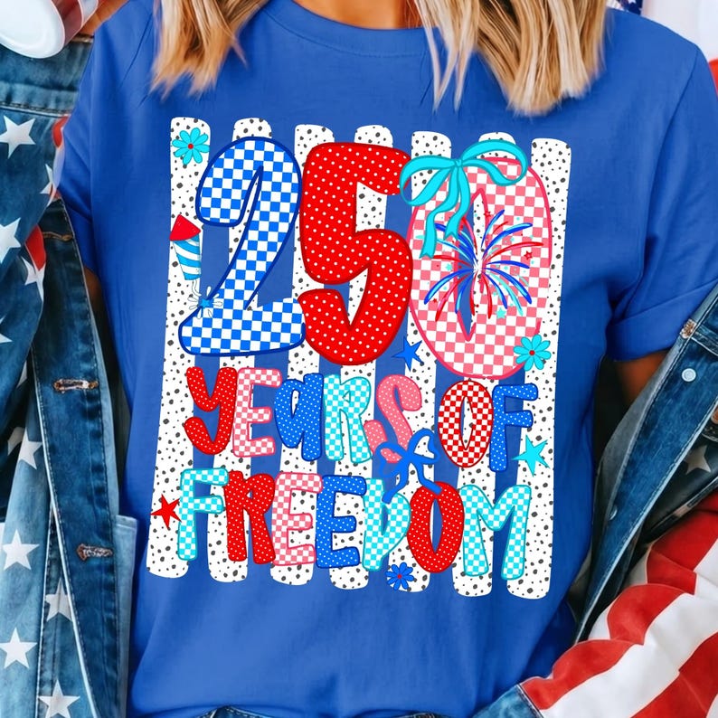 May include: A royal blue short-sleeve t-shirt featuring a graphic print with the text 250 YEARS OF FREEDOM. The design includes red, white, and blue checkered and polka-dot patterns, firework illustrations, and small blue flowers on a striped background.
