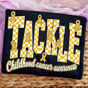 May include: A black t-shirt featuring the word TACKLE in large, yellow and white checkered block letters. Several yellow awareness ribbons are placed around the letters. Below, the text Childhood cancer awareness is printed in white script.
