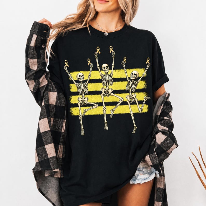 May include: A black short-sleeved t-shirt featuring a graphic of three skeletons dancing in front of three horizontal yellow brushstroke stripes. Each skeleton holds a small yellow awareness ribbon above its head in a celebratory pose.