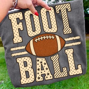 May include: A charcoal gray t-shirt featuring the word FOOTBALL in large, light-colored block letters. The letters are decorated with polka dots, stripes, and stars. A brown American football graphic is centered between the two words.