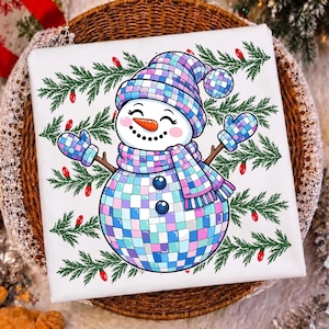 May include: A white square fabric item featuring a central graphic of a snowman made of iridescent disco ball tiles. The snowman wears a matching hat, scarf, and mittens, surrounded by green pine branches and small red Christmas light bulbs.
