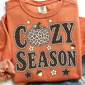 Może przedstawiać: Pomarańczowa bluza z okrągłym dekoltem z napisem COZY SEASON ciemnobrązowymi literami w białe kropki. Centralna grafika dyni ma wz&oacute;r w panterkę. Projekt zawiera małe białe kwiaty, brązowe żołędzie i gwiazdki.