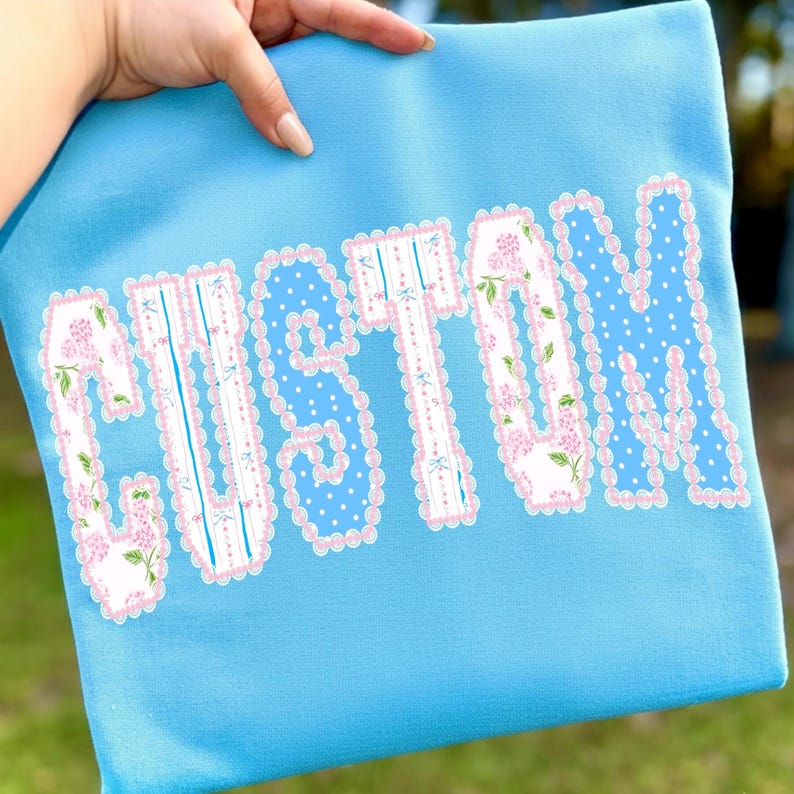 May include: A light blue fabric item with the word "CUSTOM" in large, decorative letters. The letters have a white base with pink floral and polka dot patterns, outlined with a scalloped pink border.