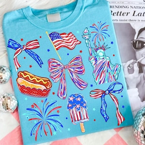 May include: A light blue t-shirt featuring a patriotic graphic print with a red, white, and blue American flag, the Statue of Liberty, a hot dog, a popsicle, fireworks, and three decorative bows in a red, white, and blue color scheme.