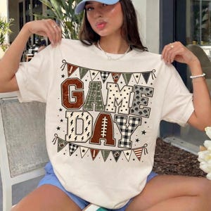 May include: A cream-colored short-sleeve t-shirt featuring the text GAME DAY in large block letters. The letters are filled with various patterns including footballs, plaid, and stars. A decorative bunting banner frames the text design.