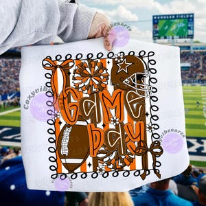 以下が含まれることがあります： 「I Love Game Day」の文字が茶色とオレンジ色で描かれた白いグラフィックTシャツ。デザインには、フットボール、ヘルメット、ポンポンが含まれています。Tシャツは黒い縁取りとオレンジと白のストライプが特徴です。