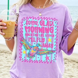 Pu&ograve; includere: Una t-shirt viola chiaro con la grafica di un'anatra colorata in volo e il testo "SOME GLAD MORNING WHEN THIS LIFE IS OVER I'LL FLY AWAY", circondata da un bordo rosa ondulato e stelle gialle su uno sfondo a righe azzurre.