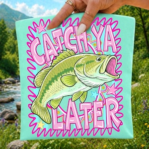 May include: A light mint green tote bag featuring a large, detailed illustration of a green bass fish in the center. The text CATCH YA LATER is printed in bold pink letters above and below the fish, surrounded by a pink squiggly border and stars.