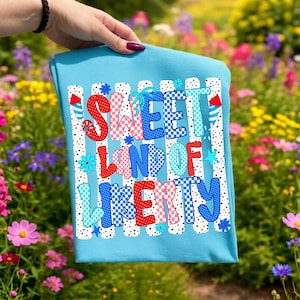 K&ouml;nnte beinhalten: Ein hellblaues T-Shirt mit dem Schriftzug "SWEET LAND OF LIBERTY" in einer kr&auml;ftigen, mehrfarbigen Schriftart mit roten, wei&szlig;en und blauen Mustern wie Punkten und Karos, akzentuiert durch kleine Feuerwerks- und Blumengrafiken.