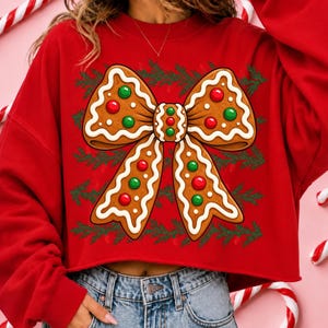 Op de afbeelding: Een felrood sweatshirt met lange mouwen voorzien van een grote afbeelding van een strik in peperkoekstijl, versierd met wit glazuur en rode en groene ronde snoepjes, op een achtergrond van kleine groene dennentakken.