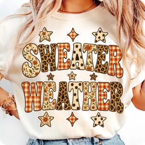 May include: A cream-colored t-shirt featuring the text "SWEATER WEATHER" in large, bold letters. Each letter is filled with a variety of patterns including orange plaid, leopard print, polka dots, and floral designs, accented by small star shapes.