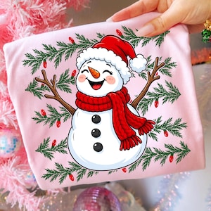 Puede incluir: Una camiseta rosa claro con el gr&aacute;fico de un mu&ntilde;eco de nieve sonriente que lleva un gorro de Pap&aacute; Noel rojo y una bufanda de punto roja. El mu&ntilde;eco tiene una nariz de zanahoria y tres botones negros, rodeado de ramas de pino y luces rojas.