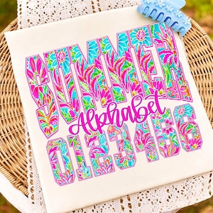K&ouml;nnte beinhalten: Cremefarbenes T-Shirt mit dem Wort "SUMMER" in gro&szlig;en, farbenfrohen Buchstaben. Die Buchstaben sind mit einem Blumenmuster in Rosa, Blau, Gr&uuml;n und Gelb gef&uuml;llt. Darunter sind das Wort "Alphabet" und die Zahlen 0-6 ebenfalls abgebildet.
