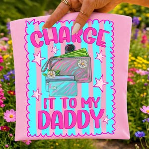 May include: A light pink fabric item featuring a bright blue and pink vertical striped background with a scalloped border. The graphic shows a wallet with green paper money and two credit cards, surrounded by stars and the text "CHARGE IT TO MY DADDY".